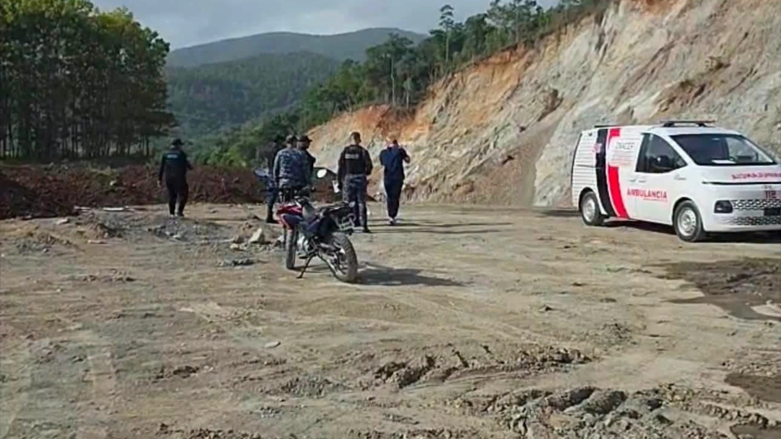 Group of people standing on a dirt road near a white ambulance with red markings beside a rocky hillside; a motorcycle sits in the foreground.