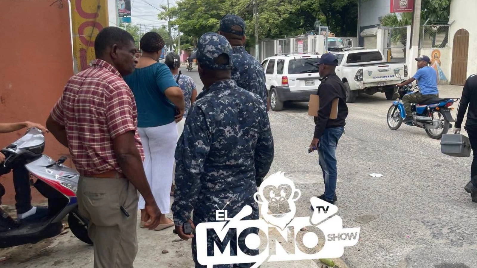 Group of civilians and uniformed officers gathered on a street corner, with a scooter and parked cars nearby, watermark foreground says 'El Mono Show'.