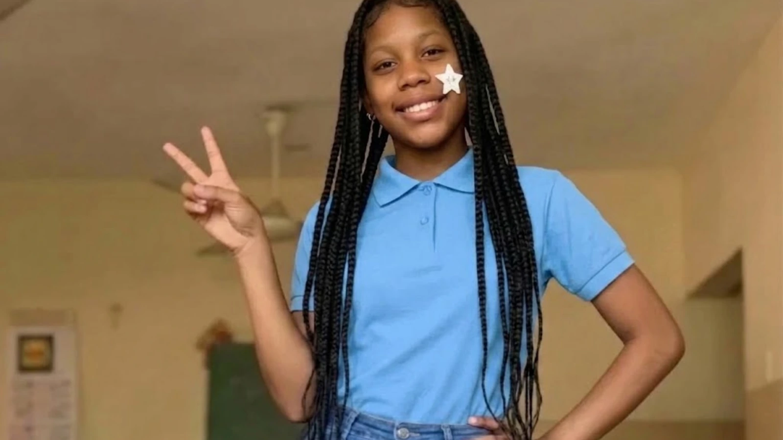 Girl smiling and posing with a peace sign in a blue polo shirt indoors, star sticker on cheek.