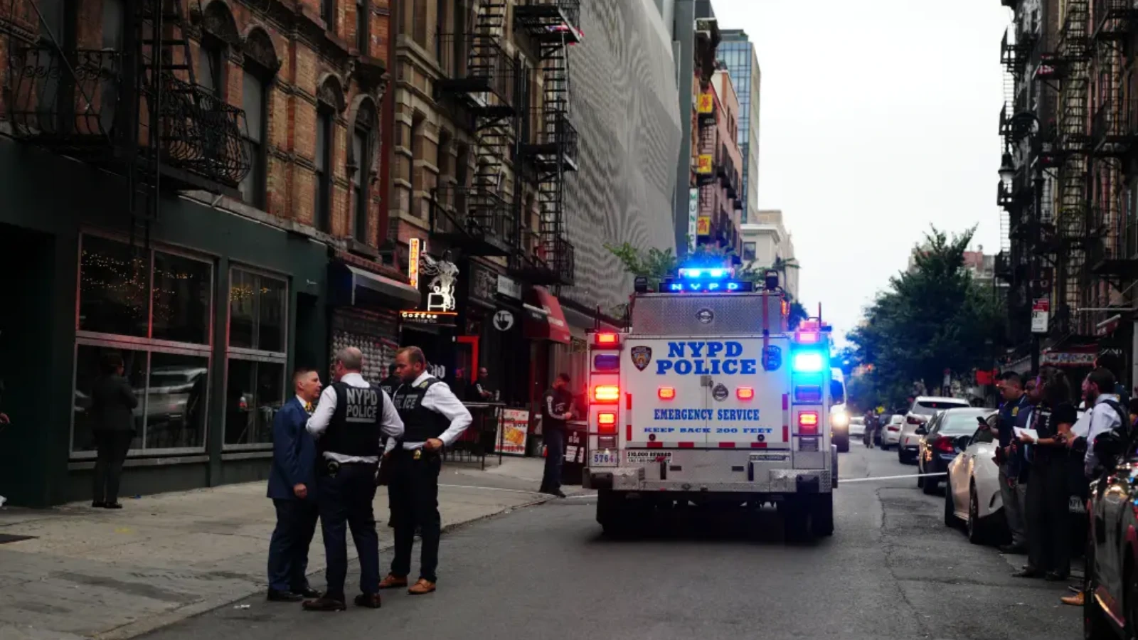 NYPD police wagon with flashing blue lights on a city street; officers talking with civilians nearby.