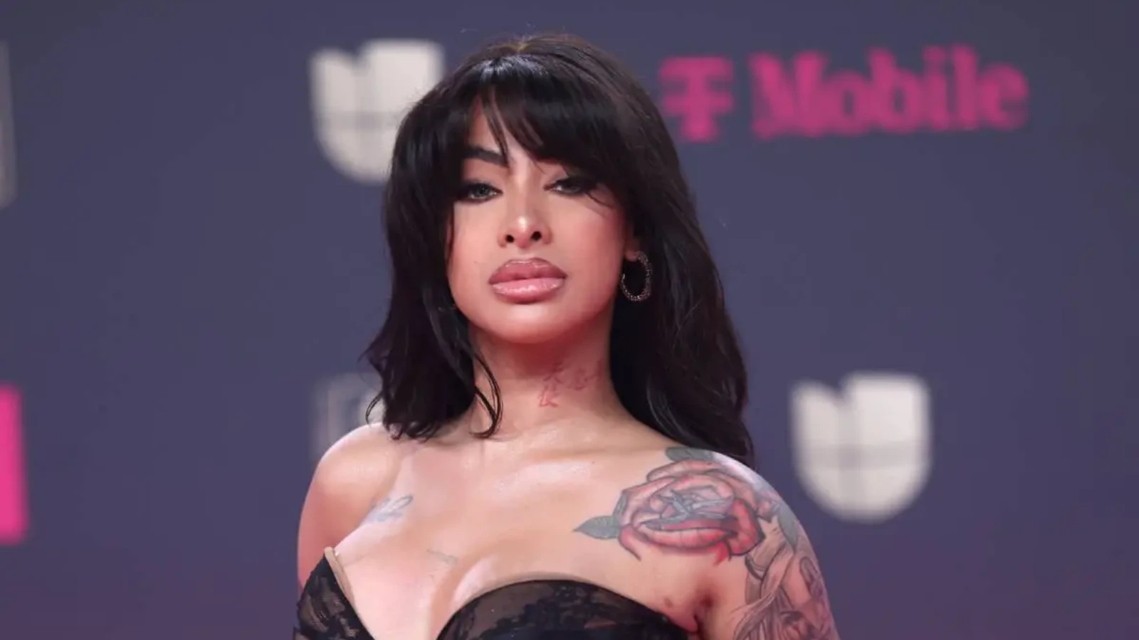 Woman with dark hair and bold makeup poses on a red-carpet backdrop, showing a rose tattoo on her left shoulder.