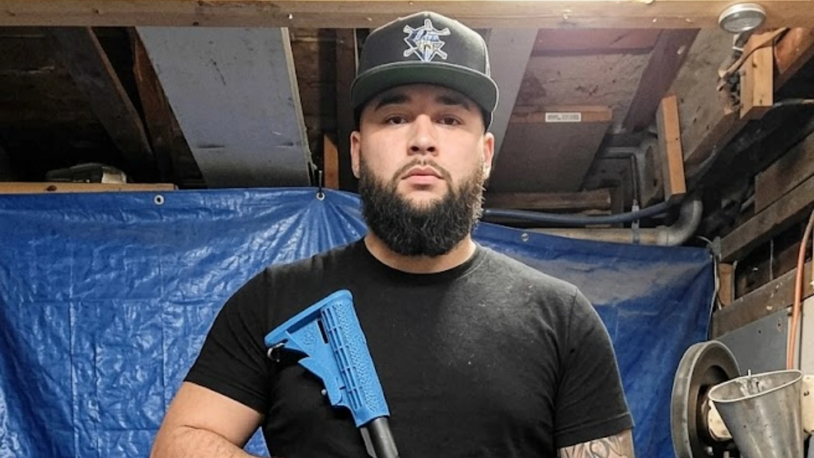Bearded man in a black t‑shirt and cap stands in a workshop, holding a blue tool.