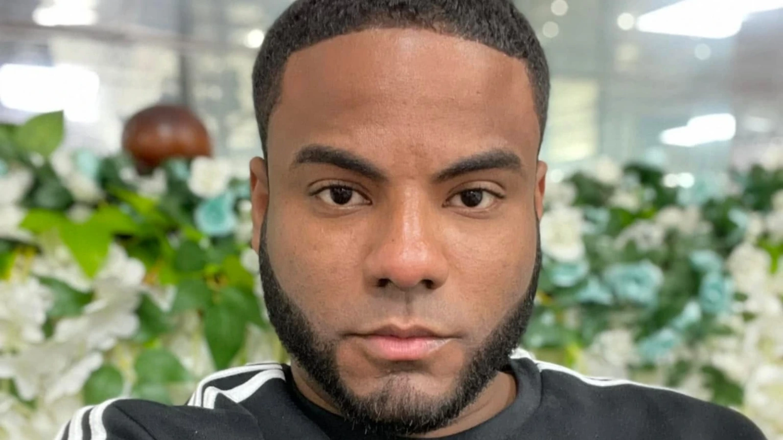Close-up portrait of a man with a short beard, looking at the camera, with a blurred floral background.