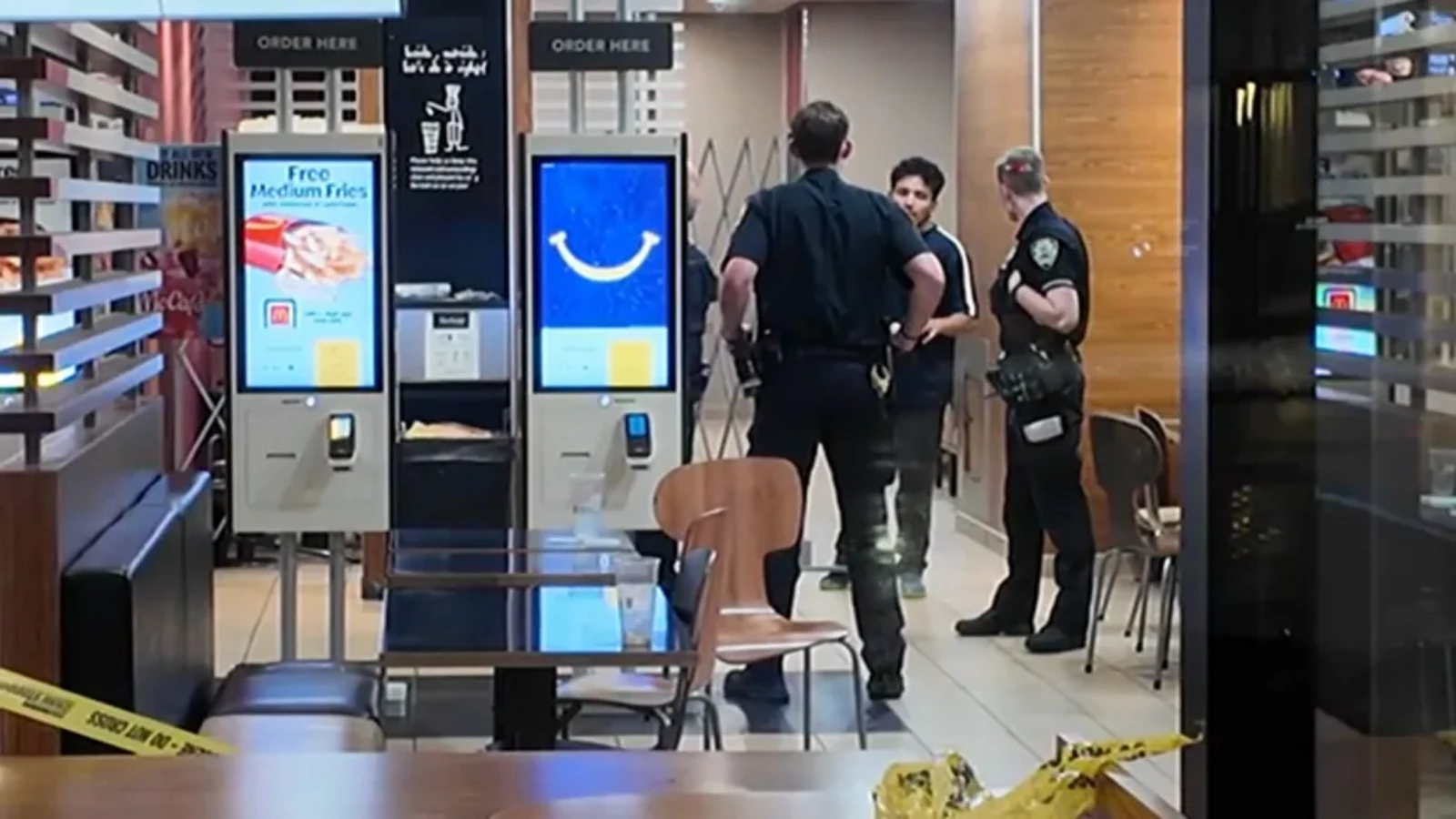 Two security officers talk with a man inside a cafe or fast-food area, yellow caution tape on the floor and multiple self-serve ordering kiosks nearby.