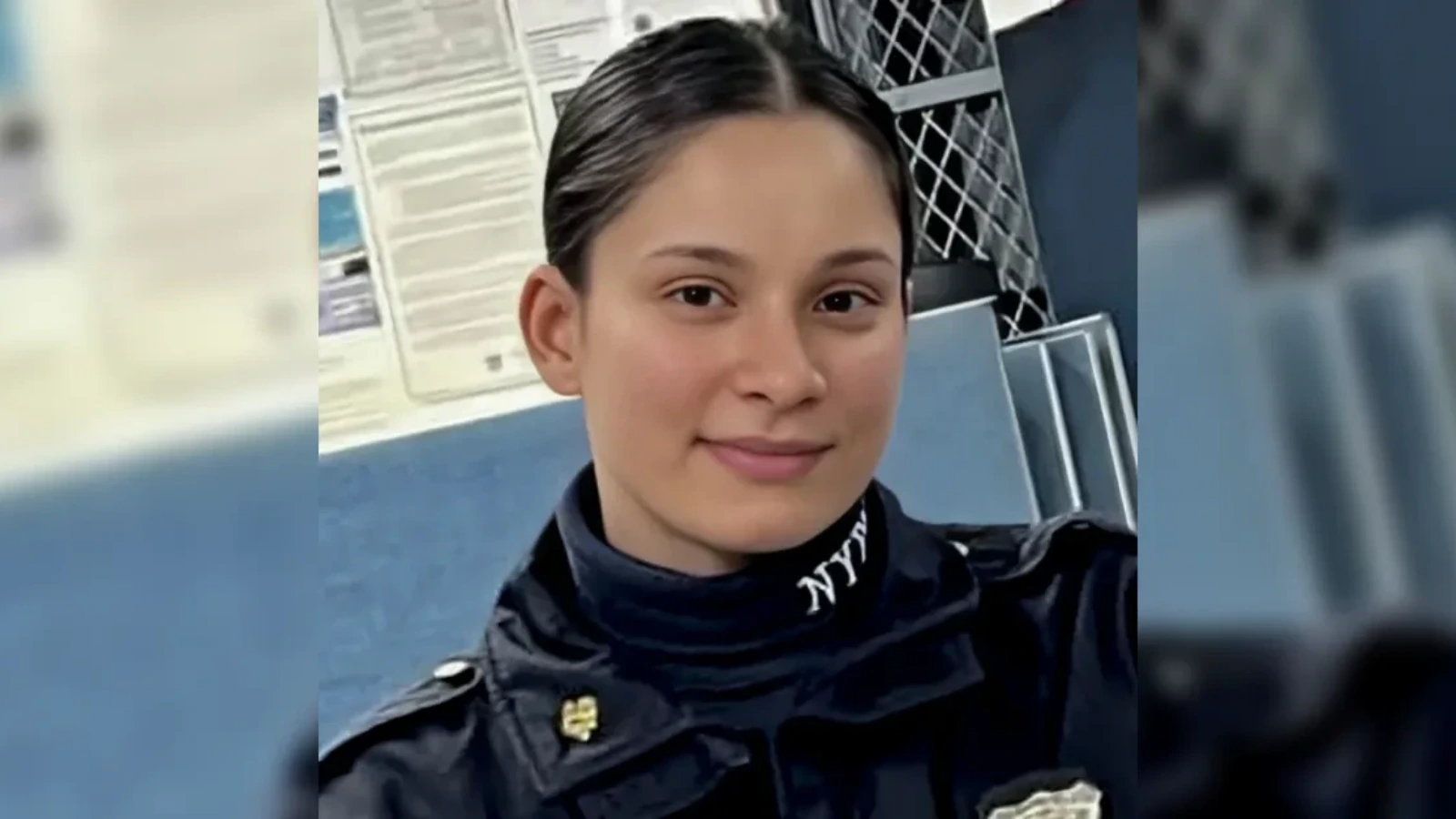 Portrait of a young woman in a dark uniform with a badge, smiling at the camera in an indoor setting, likely a security or police environment.
