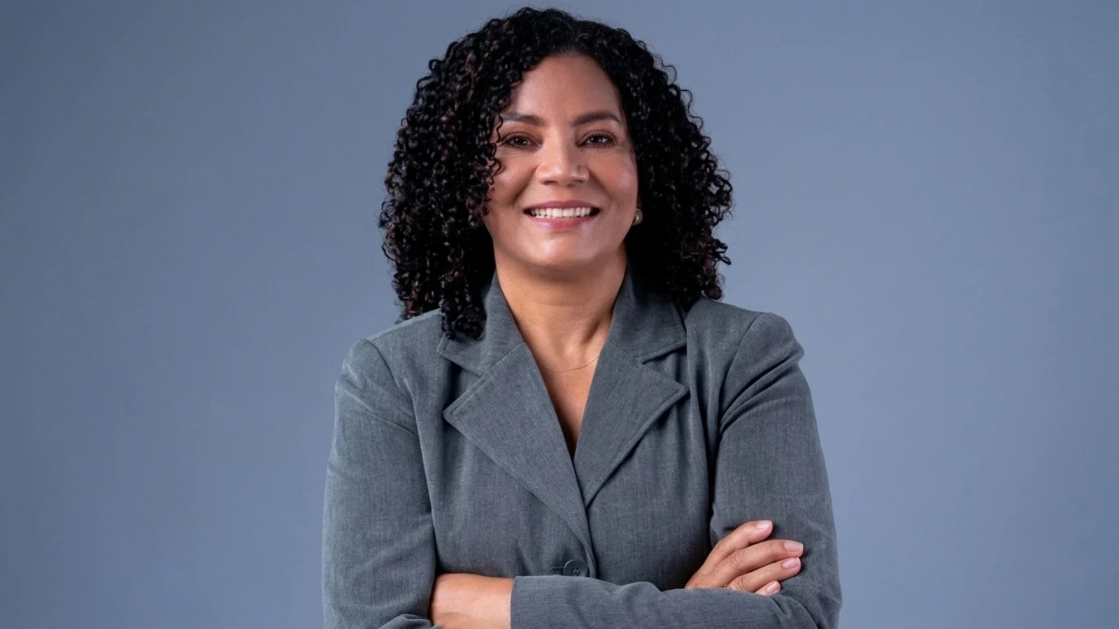 Smiling woman with curly black hair in a gray blazer, arms crossed against a blue backdrop.