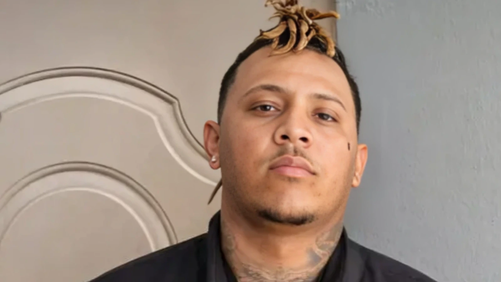 Portrait of a man with blond dreadlock top knot, facial stubble, neck tattoos, and a black shirt, standing against a gray wall.