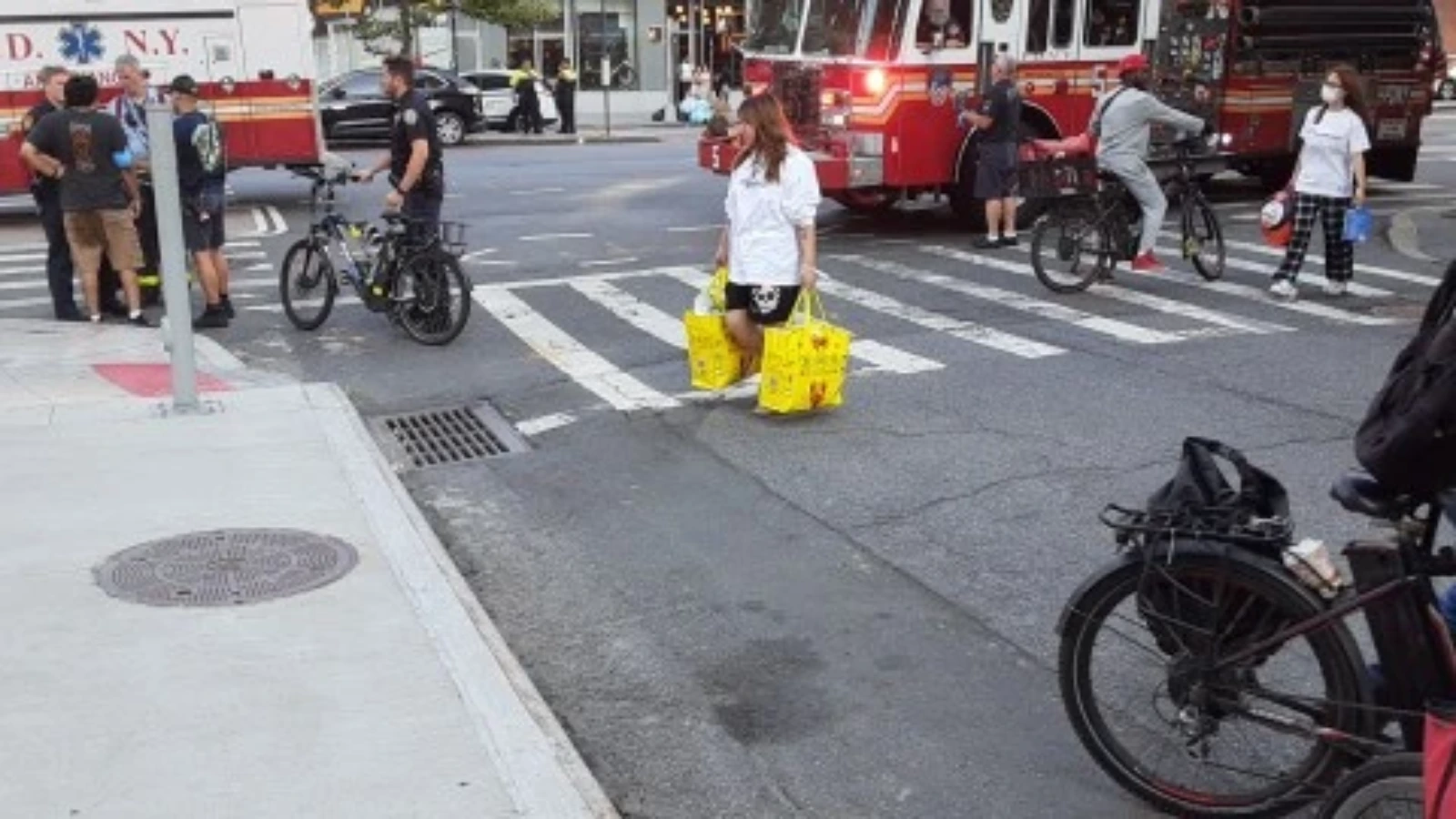 Woman in a white shirt crosses a busy street with bright yellow shopping bags; red emergency vehicles in the background.