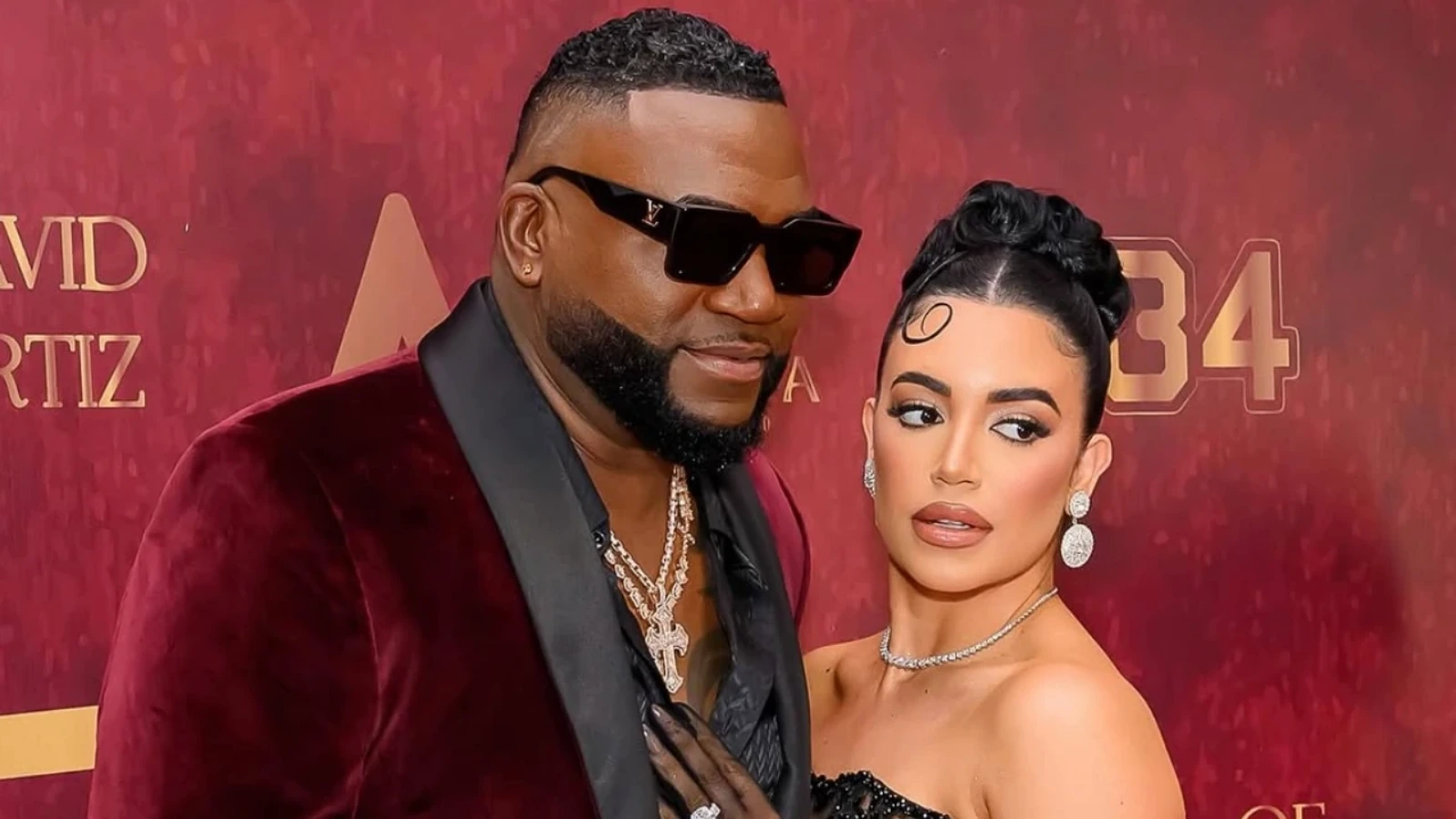 Man in a burgundy velvet suit with black lapels and chunky chains, wearing sunglasses, stands next to a woman in a black strapless dress with diamond jewelry on a red-carpet backdrop.