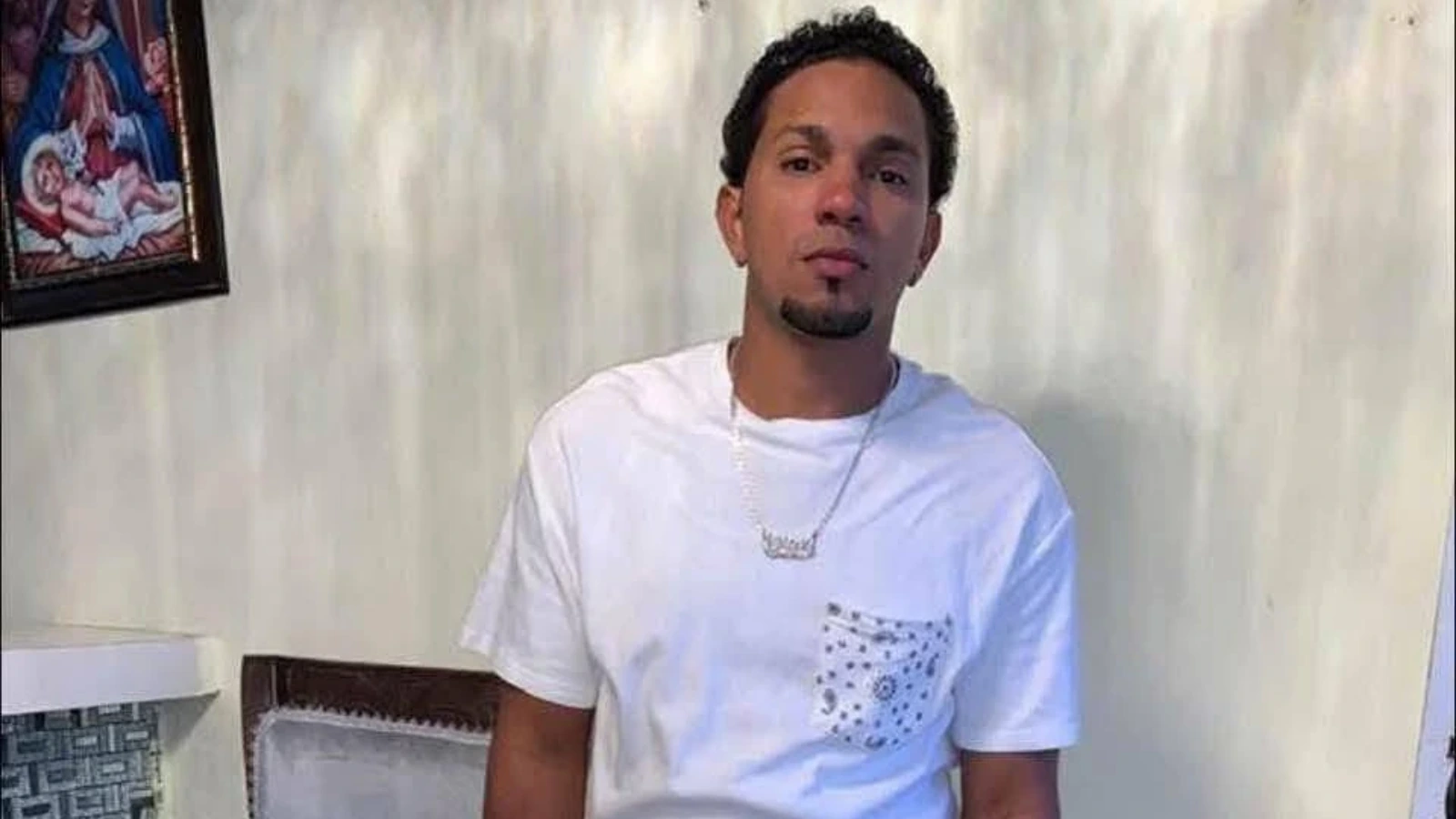 Young man wearing a white T-shirt with a patterned pocket, sitting indoors against a light wall.